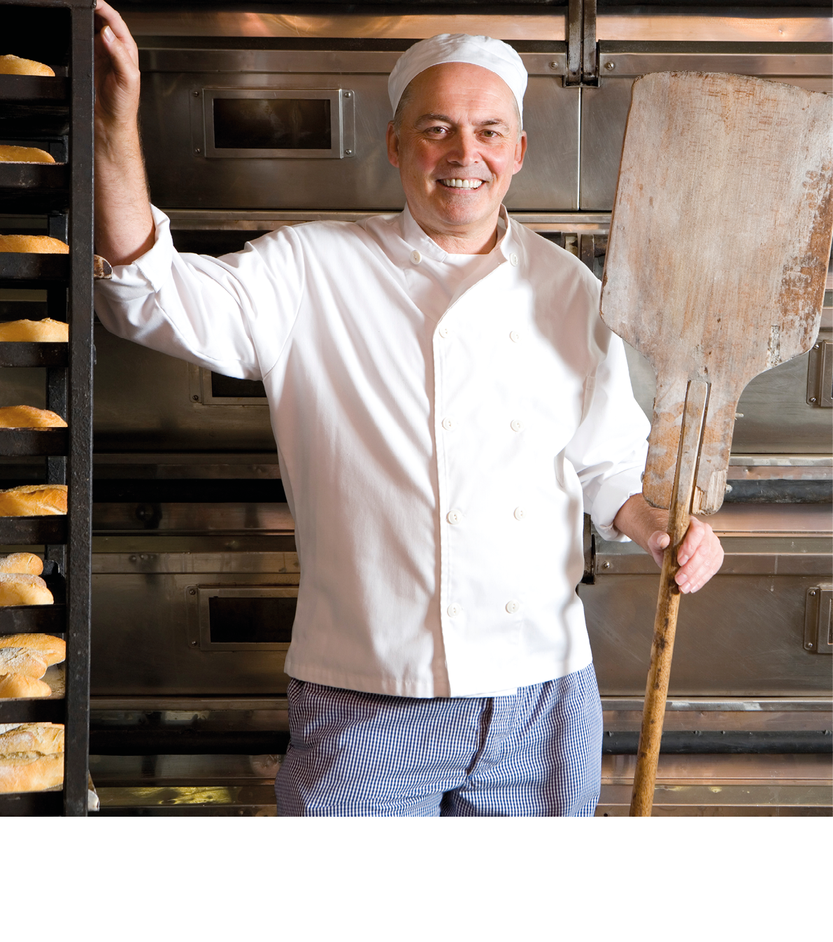 Middle-aged baker standing in his bakery with a wooden tray in his hand and smiling at the camera