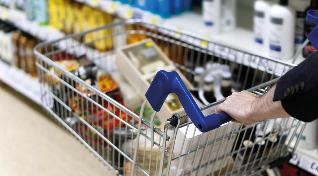 A man's arms pushing a shopping trolley full of groceries along a supermarket aisle.