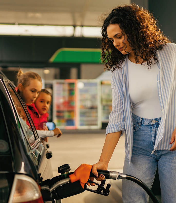 A young mother fills up gas tank at a gas station while her daughters look out the car window