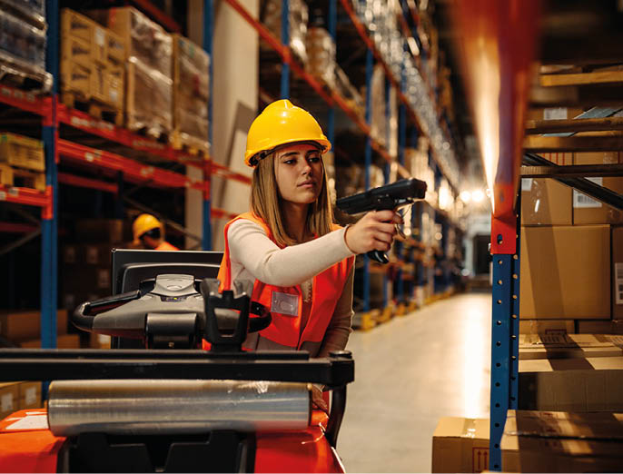 Female warehouse worker with safety helmet is working with bar code scanner while sitting on forklift in warehouse