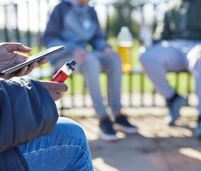 Close Up Of Teenagers With Mobile Phone Vaping and Drinking Alcohol In Park