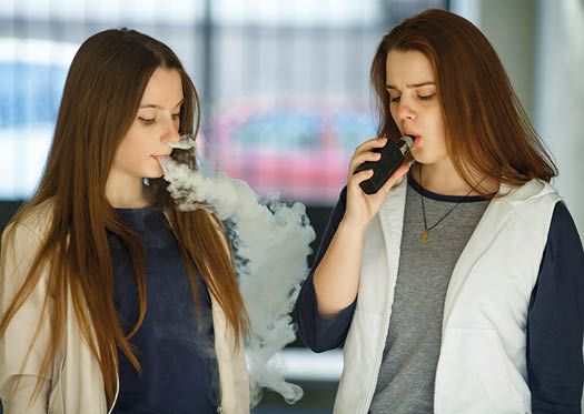 Vape teenagers. Two young cute girls in casual clothes smoke electronic cigarettes outdoors in the street in summer day. Bad habit that is harmful to health. Vaping activity.